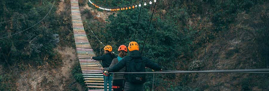 Três pessoas usando capacetes atravessam uma ponte suspensa colorida sobre uma área de floresta, segurando-se em cordas laterais para se equilibrar na Excursão a San Mateo de Otao