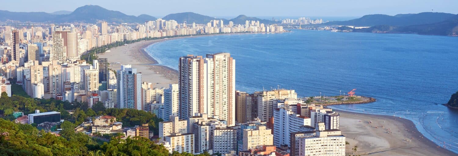 Prédios altos se alinham em um litoral curvo com uma praia de areia e montanhas ao fundo. A imagem ilustra o post sobre excursão a Santos, São Vicente e Guarujá.