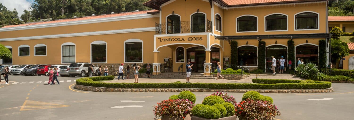 As pessoas caminham do lado de fora da Vinícola Góes em uma excursão a São Roque saindo de SP, admirando seus jardins e a fachada amarela em arco.