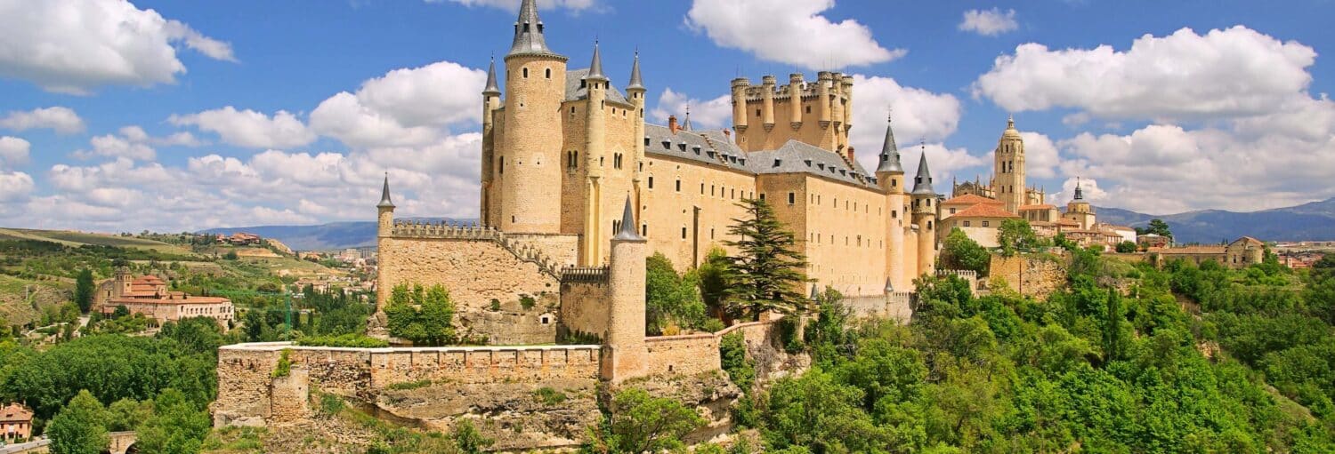 Um grande castelo de pedra com várias torres fica em uma colina cercada de vegetação sob um céu parcialmente nublado. A imagem ilustra o post sobre Segóvia, El Escorial e Vale dos Caídos.