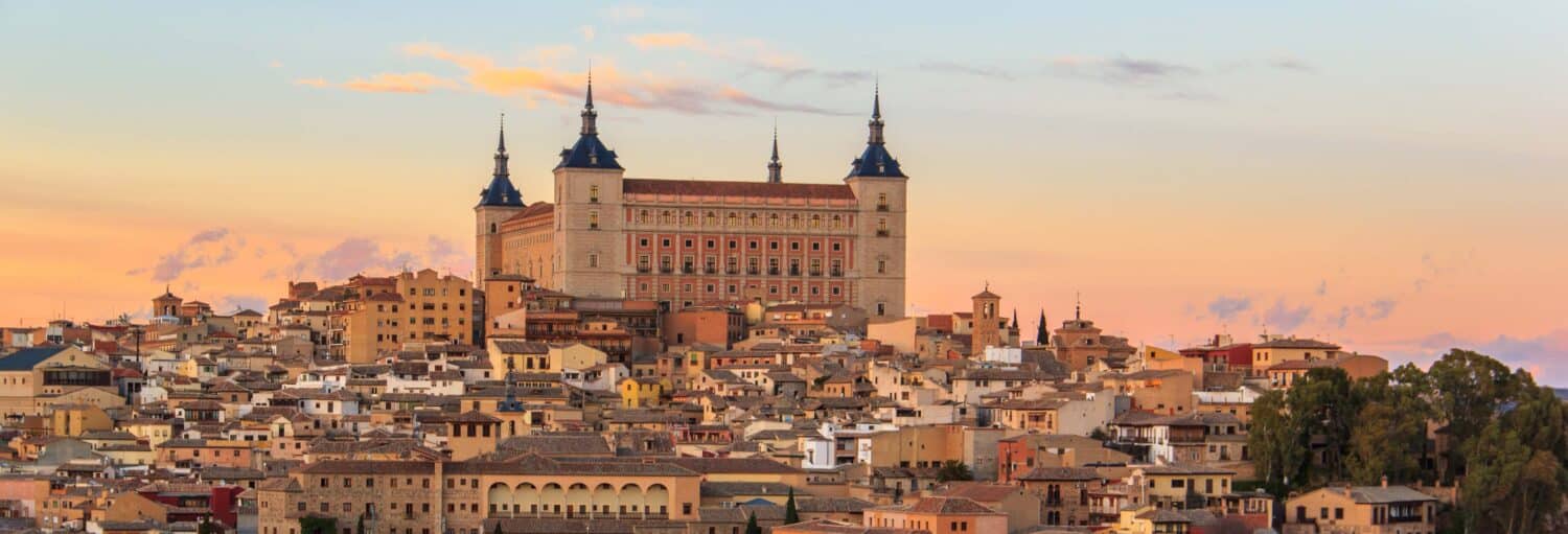 Grande edifício histórico com torres com vista para uma cidade de casas apertadas ao pôr do sol com um céu em tons pastéis. A imagem ilustra o post sobre Toledo por conta própria de ônibus.