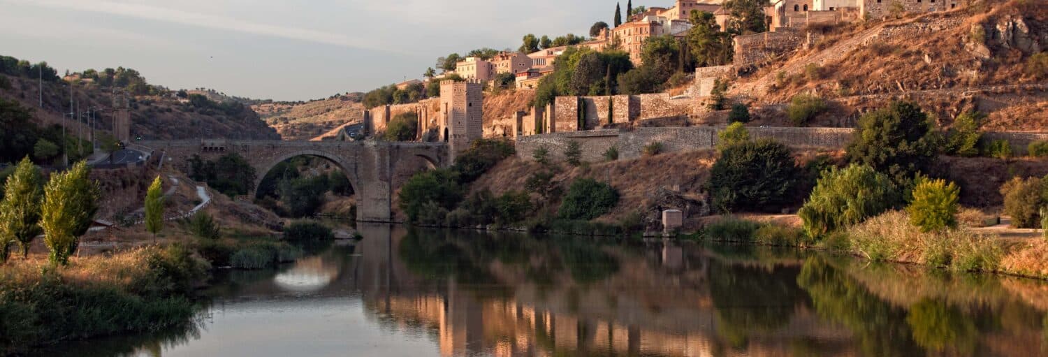 Uma ponte de pedra atravessa um rio calmo com edifícios e árvores em uma encosta ao fundo, sob um céu claro. A imagem ilustra o post sobre Excursão a Toledo + Degustação de vinhos.