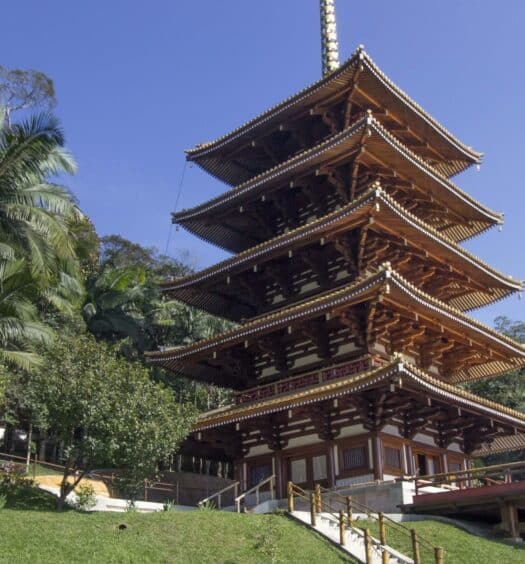 Um pagode de madeira de cinco andares coroa uma colina gramada - Excursão à Torre de Miroku saindo de SP, em meio a palmeiras e uma floresta exuberante.