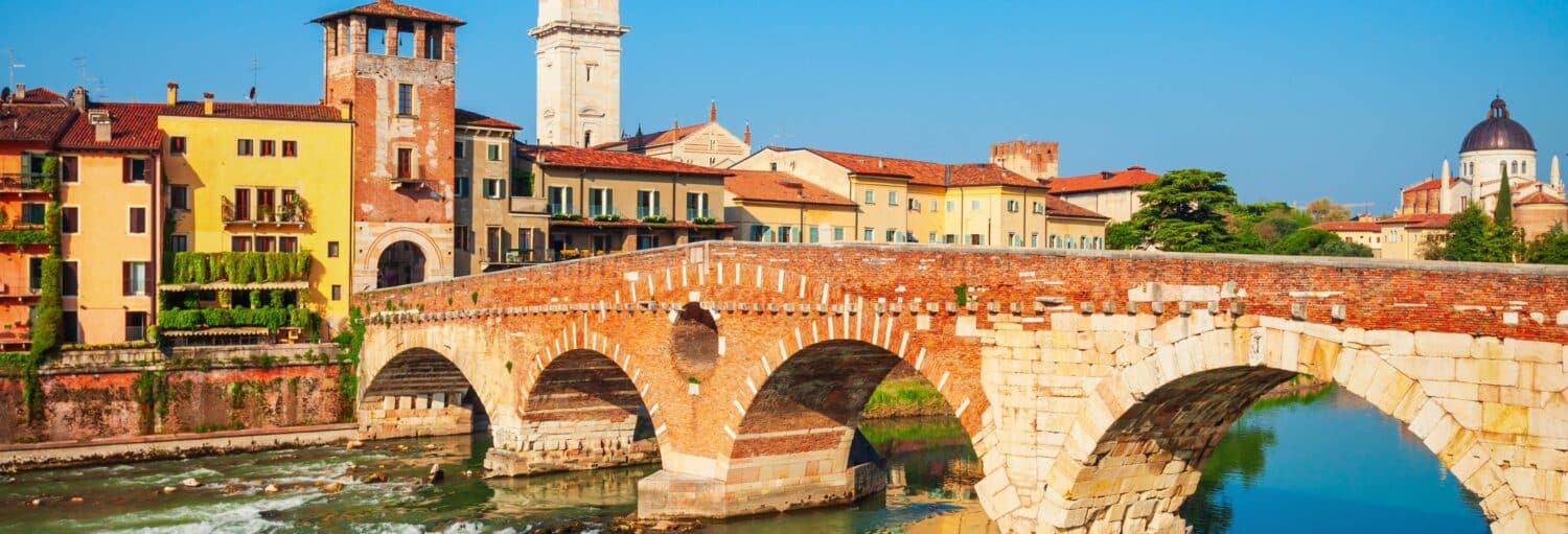 Uma ponte de pedra e tijolos em arco atravessa um rio em Verona, perfeita para uma excursão a Verona e Sirmione sob um céu azul.