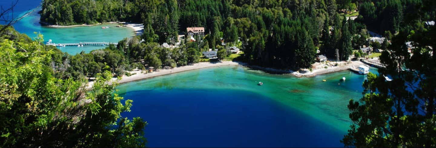 Vista aérea de uma área costeira com água azul-esverdeada clara, praias arenosas, floresta e edifícios próximos à costa na Excursão a Villa La Angostura
