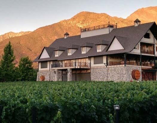 Um grande prédio de pedra da vinícola fica em frente às videiras com as montanhas ao fundo no pôr do sol. A foto é capa da Excursão à vinícola Alyan ao entardecer.