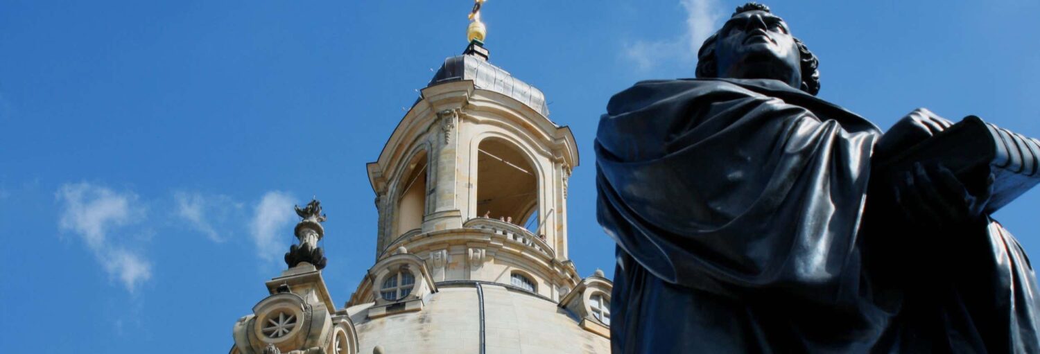 Close-up de uma estátua escura em frente à cúpula de uma igreja ornamentada contra um céu azul com algumas nuvens. Imagem para ilustrar post sobre excursão a Wittenberg.
