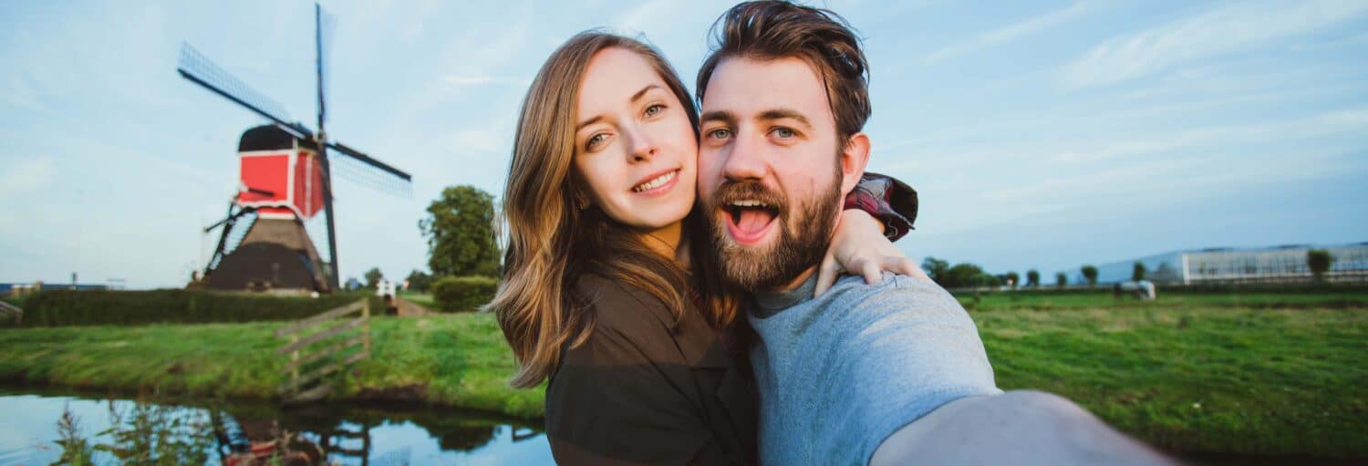 Um homem e uma mulher, ambos brancos, abraçados e tirando uma selfie em uma área rural na Holanda, com um moinho de vento no fundo - Excursão aos moinhos de vento de Zaanse Schans