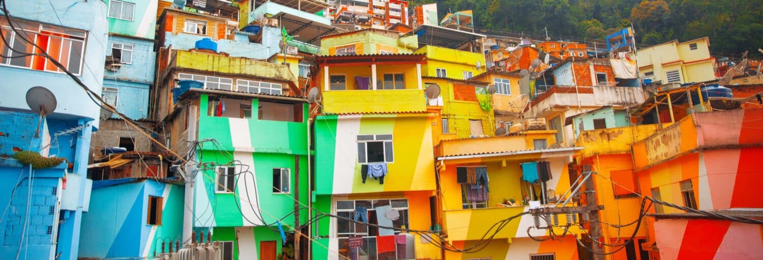 Casas coloridas estão empilhadas em uma encosta, com fios expostos e roupas penduradas do lado de fora em um bairro urbano. A imagem ilustra o post sobre Tour pela favela Santa Marta + Almoço ou Futebol.