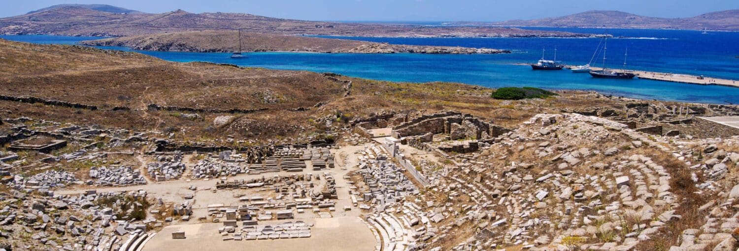 Antigas ruínas de pedra perto de um mar azul brilhante, com veleiros atracados em um píer e colinas secas ao fundo. A imagem ilustra o post sobre Ferry entre Mykonos e Delos.