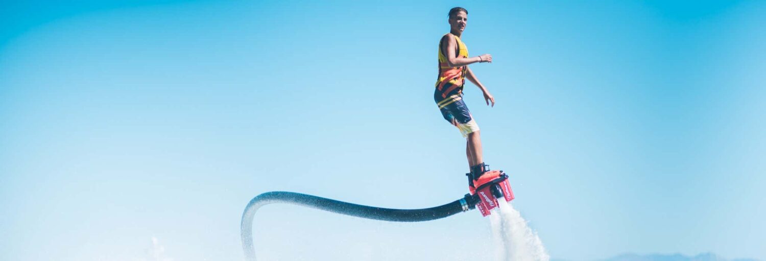 Um homem monta em um flyboard movido a água sobre a água em um céu azul claro. A imagem ilustra o post sobre Flyboard em Mykonos.