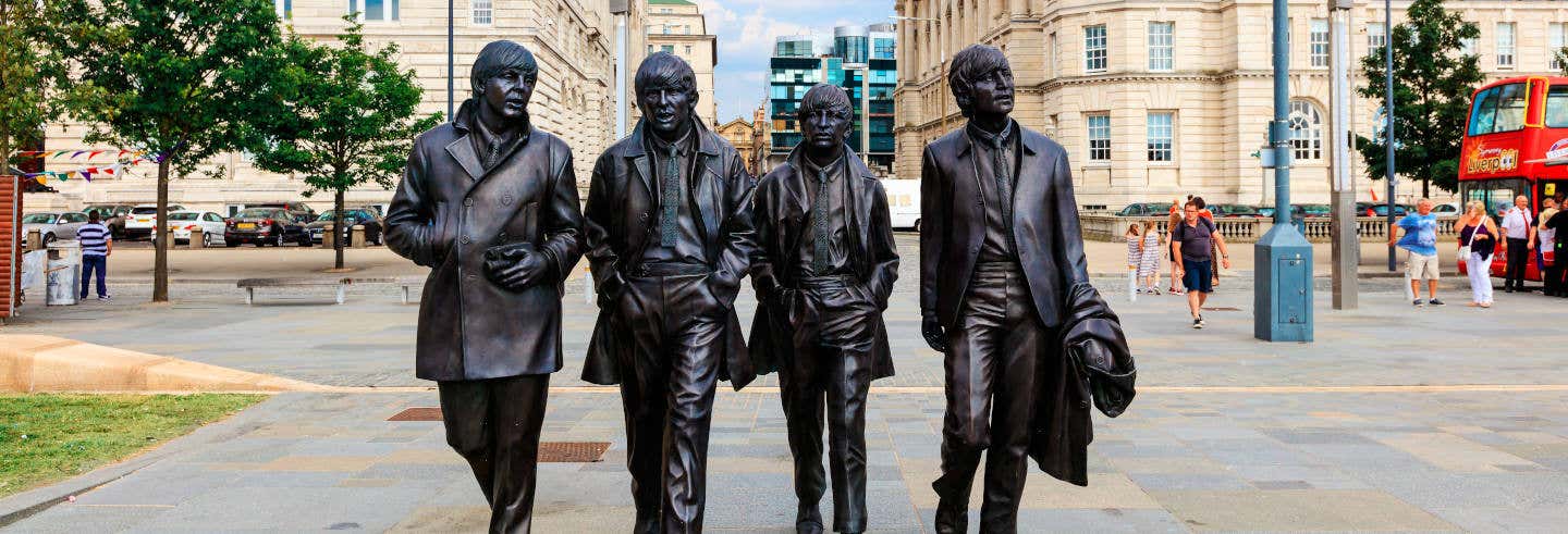 Uma estátua de bronze de quatro homens caminhando lado a lado em uma praça da cidade, com pessoas e edifícios ao fundo. A foto é capa do post do Free tour dos Beatles.