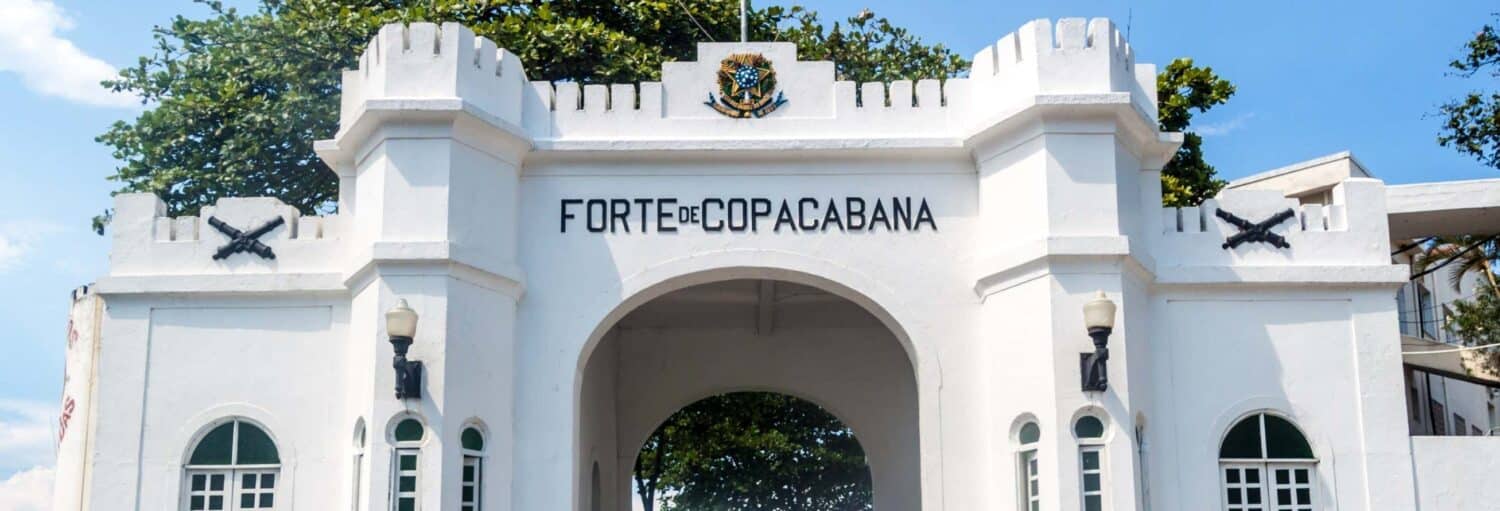 Arco de entrada branco do Forte de Copacabana com texto em preto e insígnia militar, ladeado por dois postes de iluminação. A imagem ilustra o post sobre Free tour por Copacabana e Ipanema.