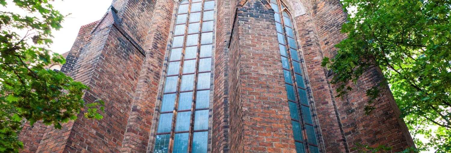 Parede alta de tijolos da igreja com janelas grandes e estreitas e galhos verdes de árvores emoldurando parcialmente o edifício. Imagem para ilustrar post sobre o free tour dos mistérios e lendas de Berlim.