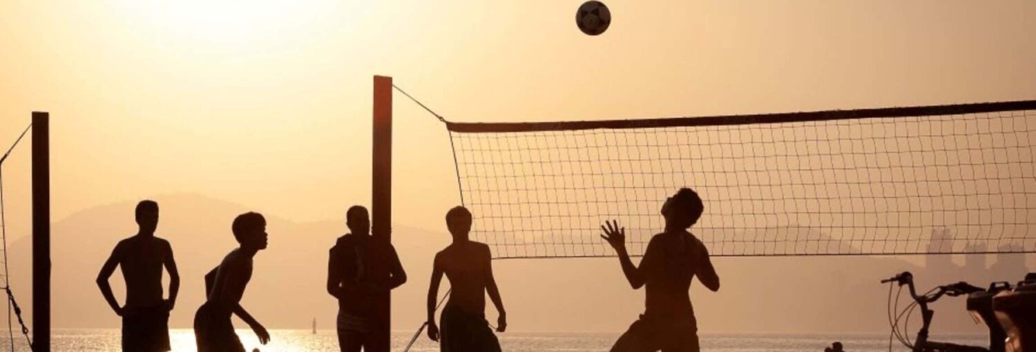 Cinco pessoas jogam futevôlei na praia de Copacabana ao pôr do sol; a bola voa acima da rede, com bicicletas por perto.