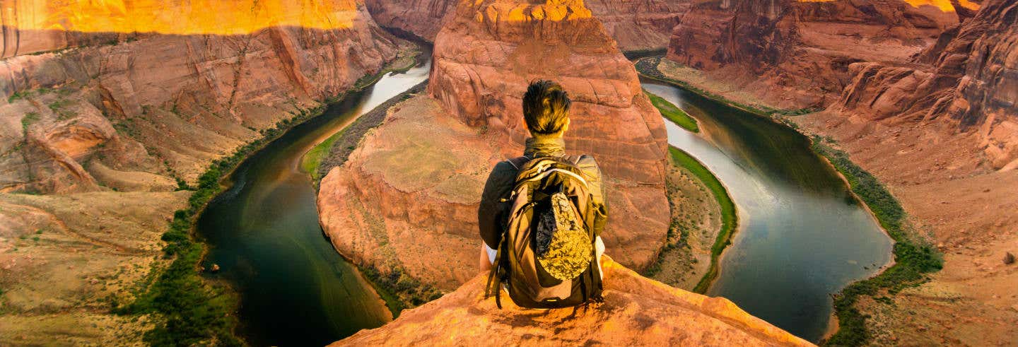 Uma pessoa com uma mochila senta-se em um penhasco com vista para uma curva de rio cercada por paredes íngremes de cânions de rocha vermelha. Imagem para ilustrar post sobre Grand Canyon e Horseshoe Bend em 2 dias.