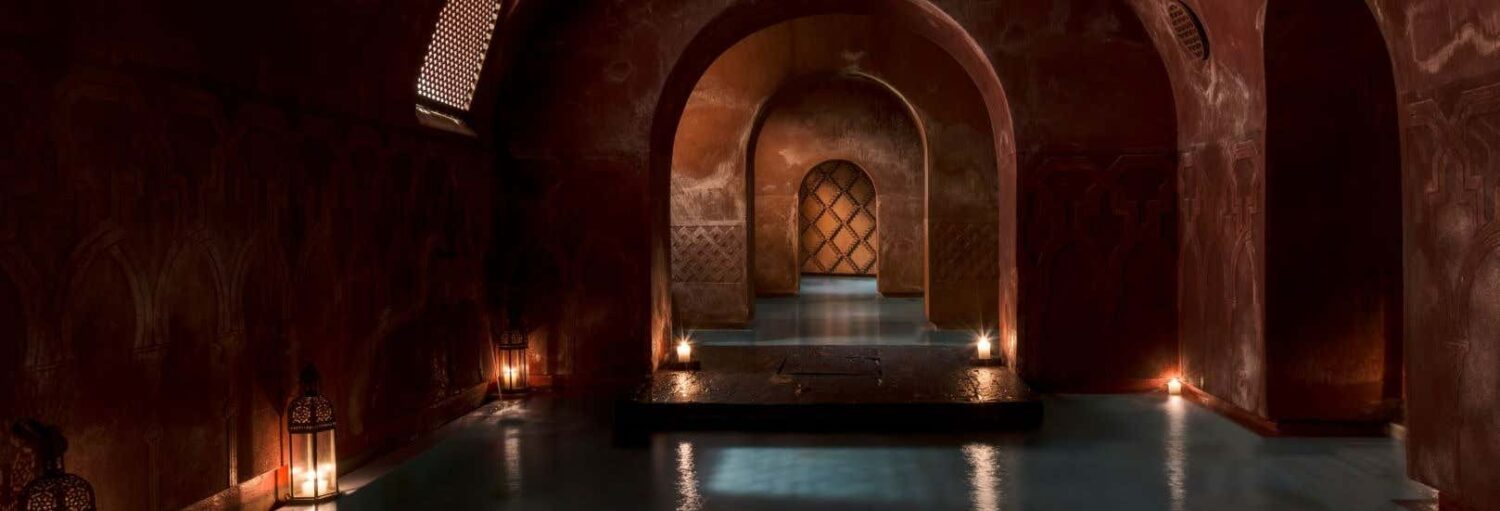Piscina interna pouco iluminada com portas em arco, lanternas e paredes estampadas em um ambiente semelhante a um spa. A imagem ilustra o post sobre Hammam Al Ándalus.