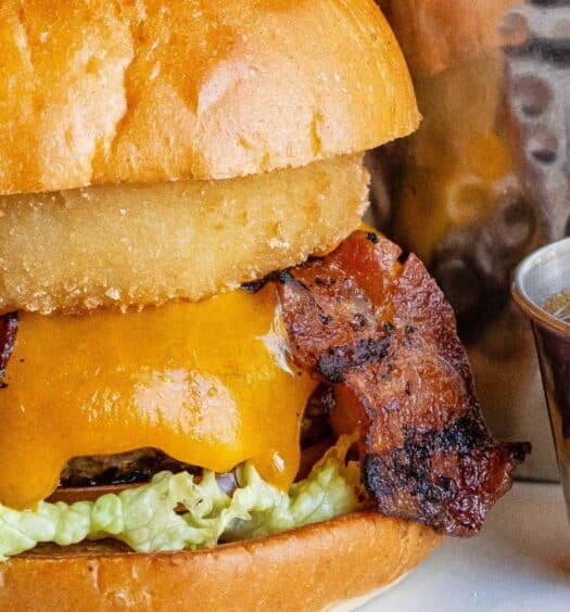 Um cheeseburger com alface, bacon, cheddar e um anel de cebola, servido com um copo metálico de molho em um prato branco. A imagem ilustra o post sobre Hard Rock Cafe Miami sem filas.