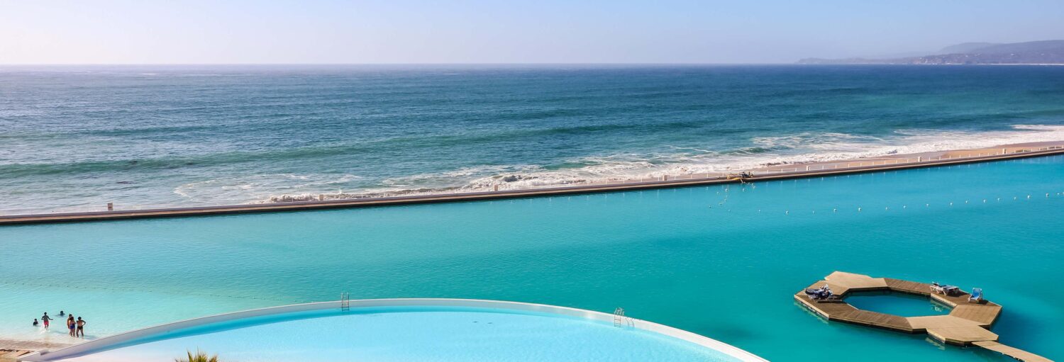Grande piscina ao lado do oceano com uma seção circular e pessoas em pé na água rasa.