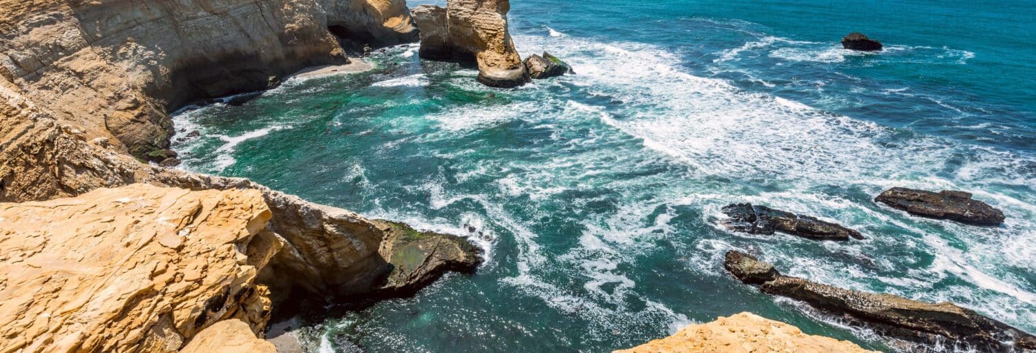 Penhascos costeiros rochosos com vista para a água azul-turquesa do oceano, com ondas batendo contra rochas e pilhas de mar nas Ilhas Ballestas + Reserva Nacional de Paracas