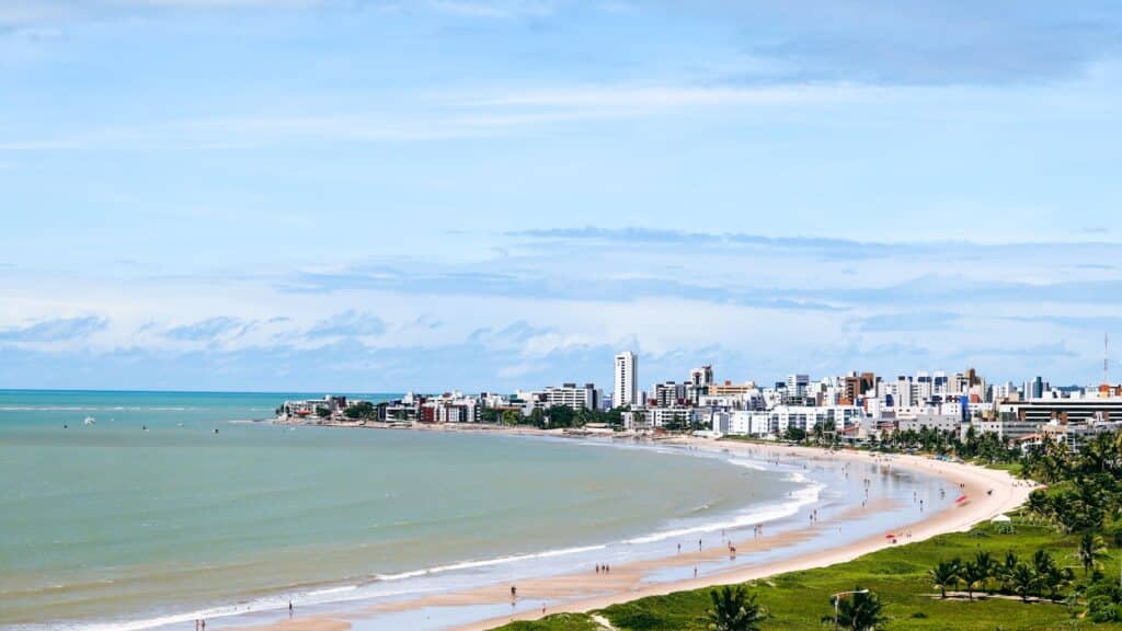 Uma paisagem urbana litorânea com prédios altos, uma praia de areia, áreas verdes e águas calmas do oceano sob um céu parcialmente nublado, em João Pessoa.