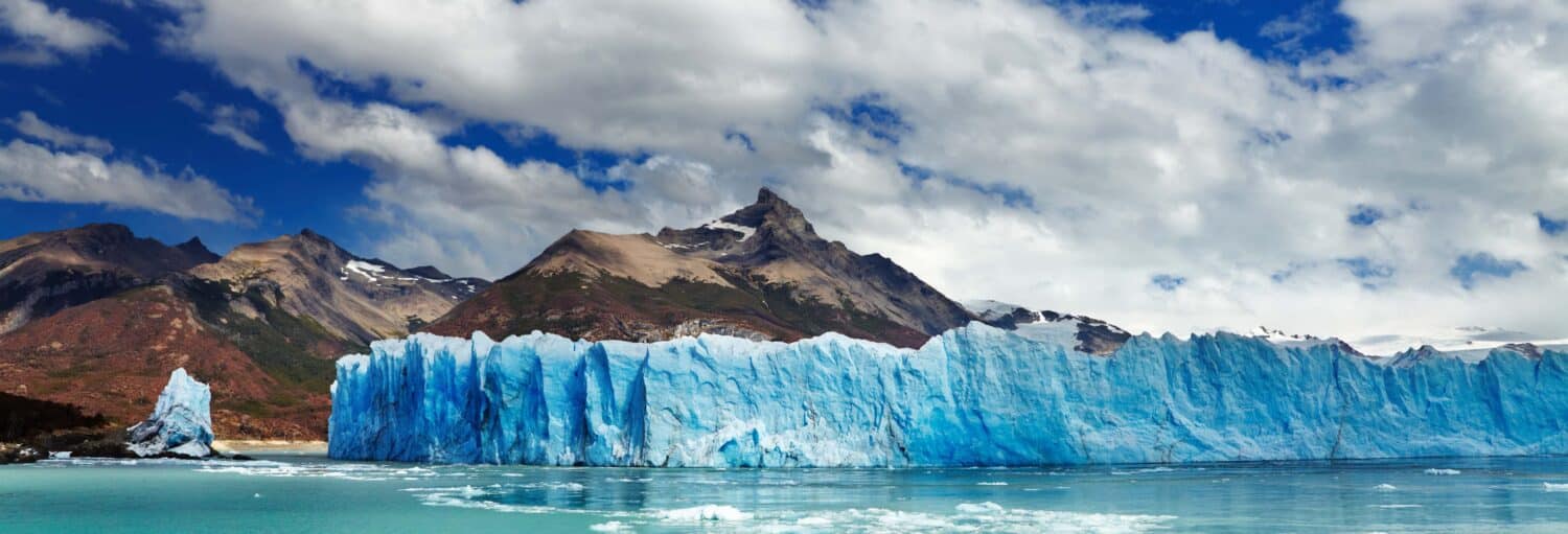 Uma grande geleira azul se estende pela água com montanhas e um céu parcialmente nublado ao fundo. Imagem para ilustrar post sobre o tour de caiaque pelo glaciar Perito Moreno.