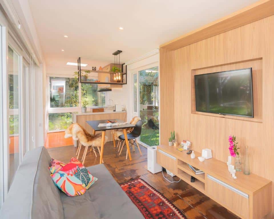 Moderna sala de estar e cozinha em plano aberto com detalhes em madeira, TV na parede, mesa de jantar e grandes janelas para um jardim, na KŌN - Pousada Romântica.