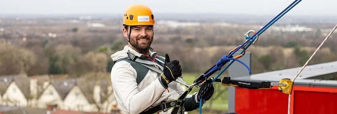 Um homem com equipamento de segurança e capacete sorri e faz sinal de positivo enquanto faz rapel em um prédio com cordas. A foto é capa do post do Ingresso do Museu do Liverpool FC + Rapel em Anfield.