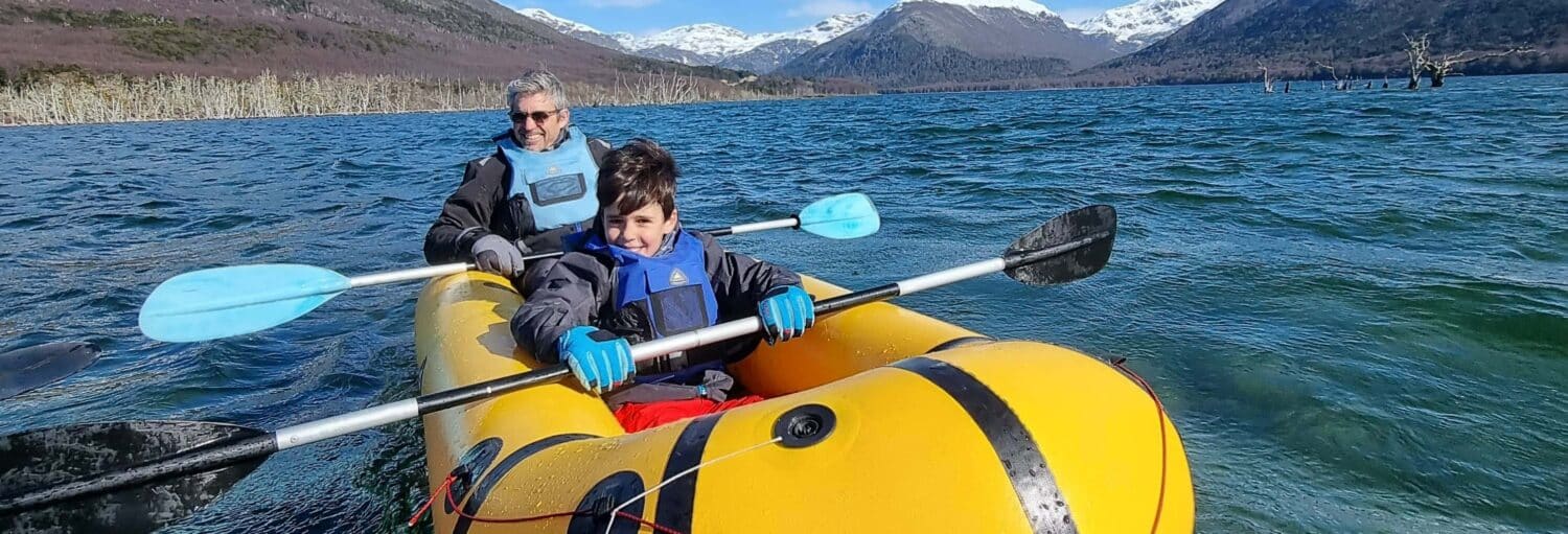 Duas pessoas usando coletes salva-vidas remam em um caiaque inflável amarelo em um lago com montanhas ao fundo. Imagem para ilustrar post sobre packrafting de 2 dias pelo rio Lasifashaj.