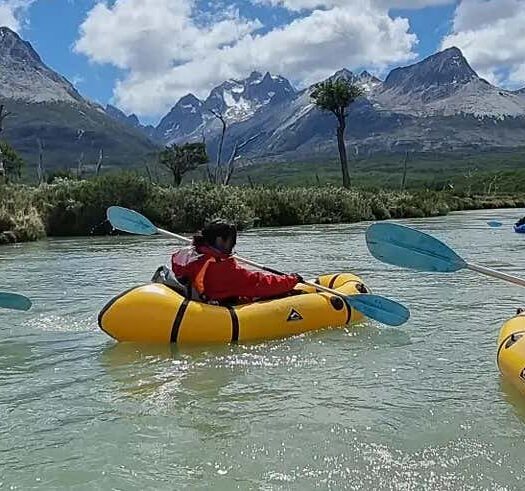 Três pessoas em caiaques infláveis remam por um rio com montanhas e árvores ao fundo sob um céu parcialmente nublado. Imagem para ilustrar post sobre packrafting no rio Olivia.