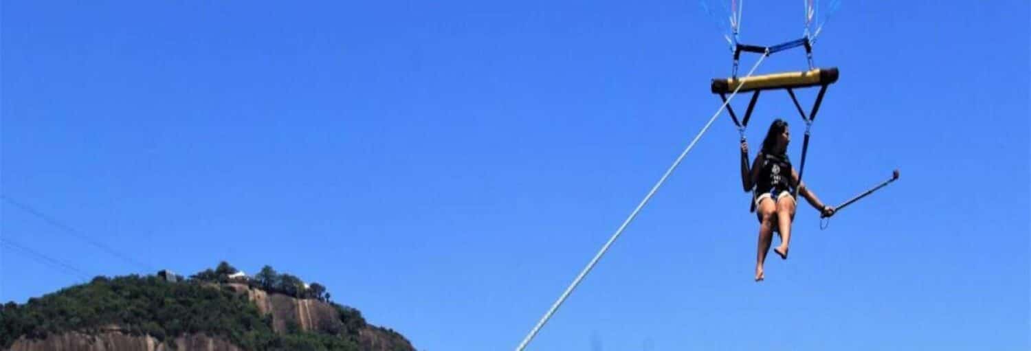 Uma pessoa está fazendo parasailing no Rio de Janeiro, segurando um bastão de selfie no alto de uma colina contra um céu azul claro.