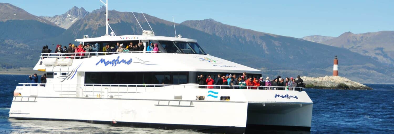 Um catamarã branco com muitos passageiros navega em um lago próximo a montanhas e um pequeno farol em uma ilhota rochosa. Imagem para ilustrar post sobre o passeio de barco pelo Canal Beagle.