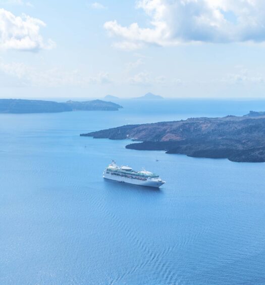 Um grande navio de cruzeiro navega em águas calmas e azuis perto de uma ilha sob um céu parcialmente nublado. A imagem ilustra o post sobre Barco com fundo de vidro pela Caldeira de Santorini.