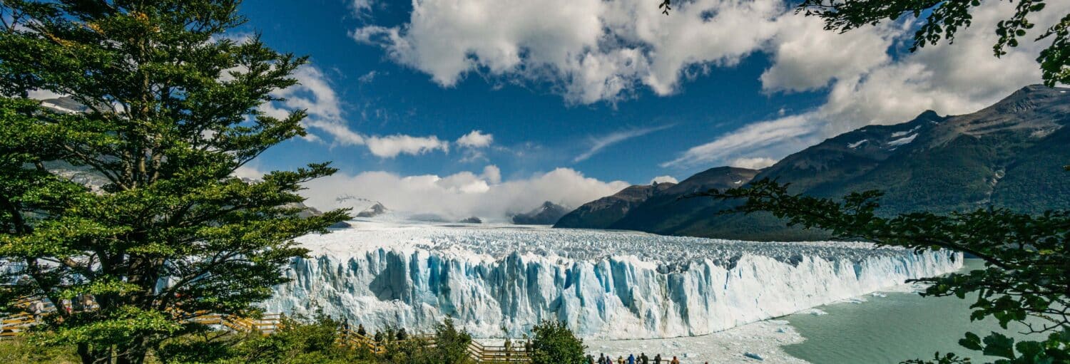 Grande geleira com bordas de gelo irregulares, cercada por montanhas, árvores, céu azul e nuvens dispersas. Pessoas em pé em um deck de observação. Imagem para ilustrar post sobre passeio de barco pelo Lago Argentino.