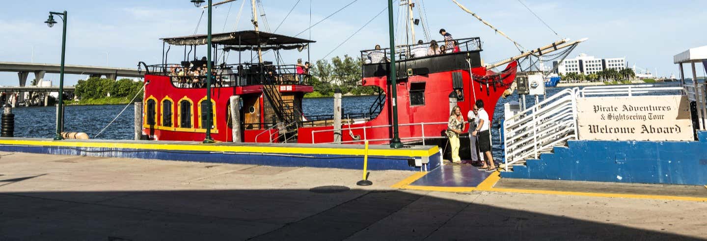 Um barco vermelho e preto com tema de pirataria está ancorado perto de uma passarela com uma placa de "Bem-vindo a bordo" e pessoas próximas. A imagem ilustra o post sobre Passeio de barco pirata por Miami.