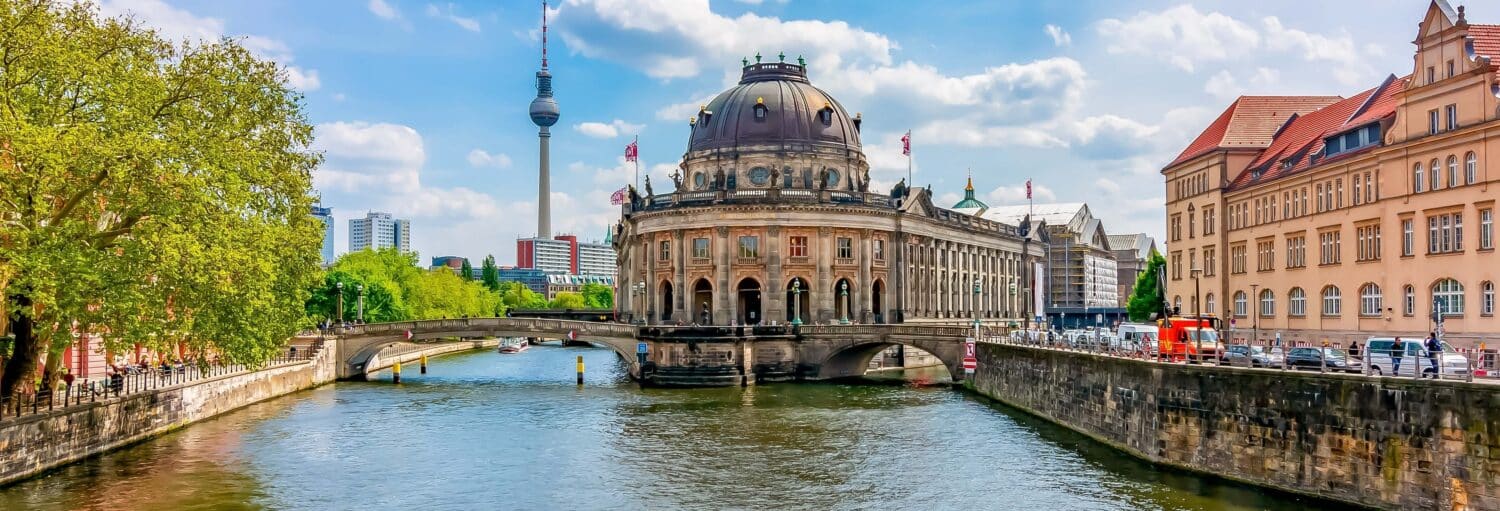 Um edifício abobadado fica em uma ilha em um rio, com uma torre de TV e edifícios da cidade ao fundo, sob um céu azul. Imagem para ilustrar post sobre passeio de barco pelo rio Spree.
