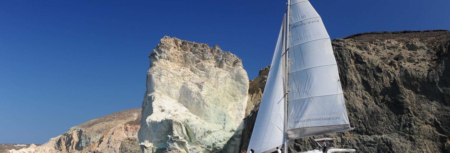 Um catamarã privado pela Caldeira de Santorini navega perto de falésias sob um céu azul e formações rochosas imponentes.