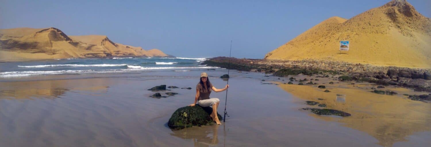 Uma pessoa sentada em uma pedra segurando uma vara de pescar em uma praia de areia com dunas e o oceano ao fundo na Pesca no sul de Lima