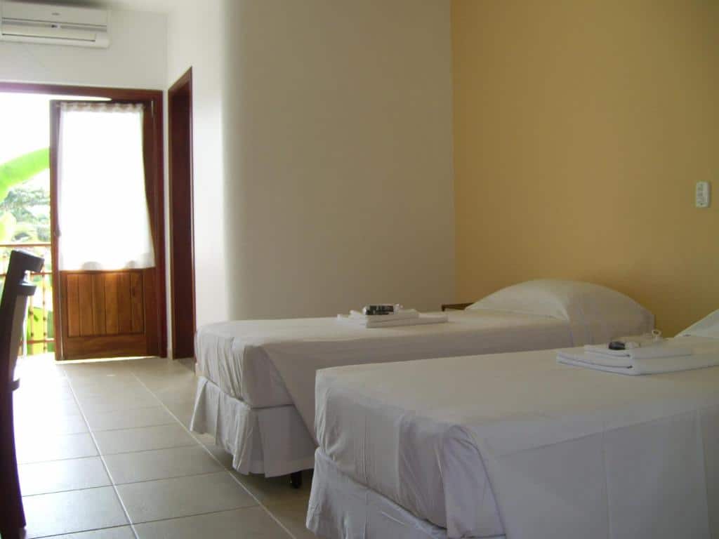 Duas camas de solteiro com roupa de cama branca em um quarto simples; porta aberta para o exterior, permitindo a entrada de luz natural, em um quarto na Pousada do Tié na Praia do Itaguá.