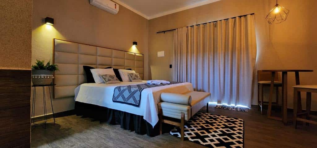 Um quarto moderno na Pousada Lago Azul em Delfinópolis com uma cama de casal, luzes de cabeceira, uma pequena mesa com banquetas, cortinas e um tapete geométrico.