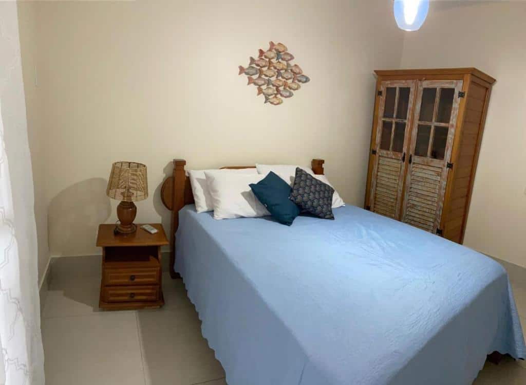 Um quarto no Porto Bali Apartamentos com cama de madeira, colcha azul, criado-mudo com abajur e um guarda-roupa de madeira contra uma parede creme.