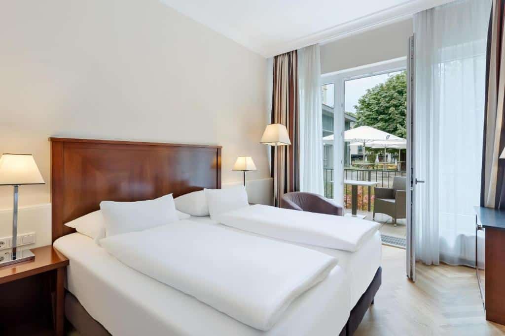 Um quarto no Austria Trend Parkhotel Schönbrunn Wien com uma cama de casal, duas luminárias, uma cadeira e uma porta aberta que leva a uma varanda com assentos ao ar livre, para representar onde ficar em Viena.