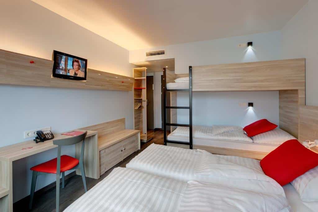 Um quarto no Meininger Hotel Wien Downtown Franz com um beliche, uma cama de solteiro, uma escrivaninha, uma TV e travesseiros vermelhos, para reprensentar onde ficar em Viena.