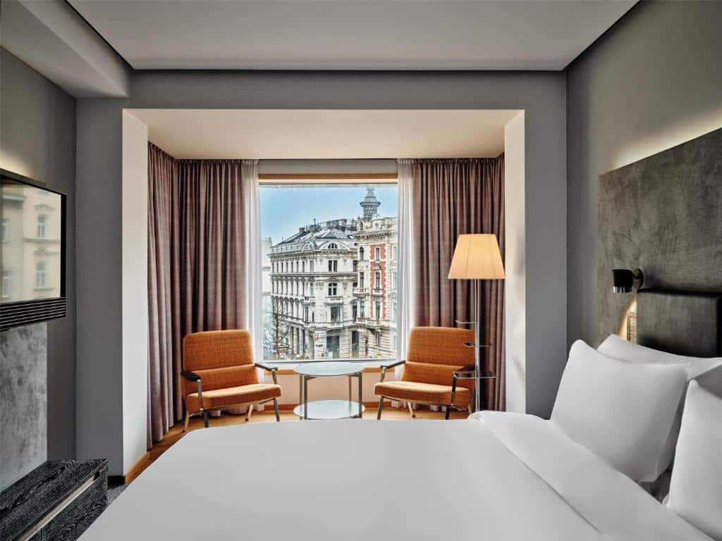 Quarto no Radisson Blu Das Triest Hotel com uma cama grande, duas poltronas perto de uma janela, uma luminária de chão e vista externa para um prédio da cidade, para representar onde ficar em Viena.