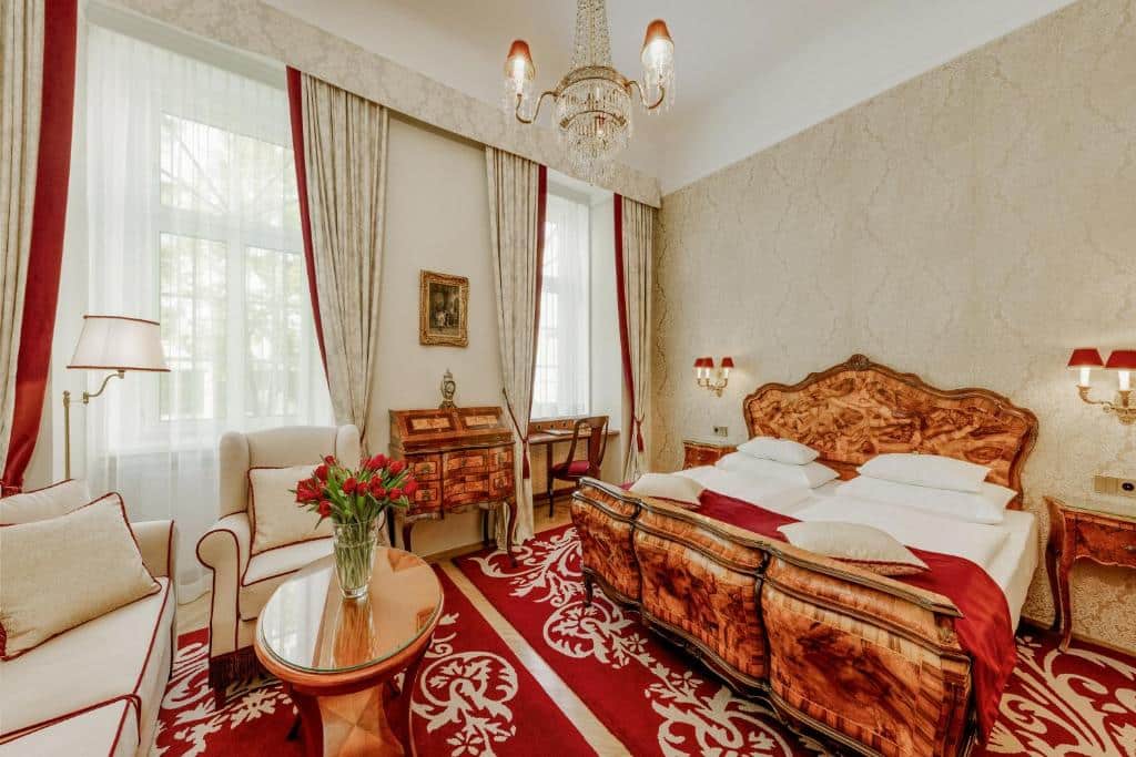 Quarto no Hotel Stefanie com móveis de madeira ornamentados, detalhes em vermelho, cama de casal, sofá, escrivaninha e janelas grandes com cortinas.