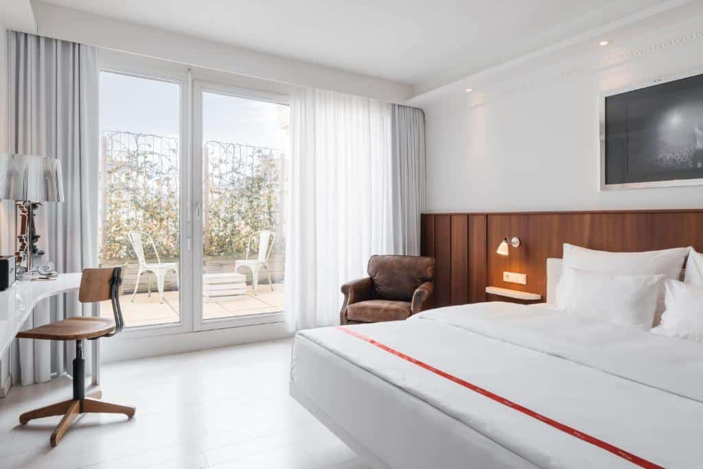 Um quarto no Ruby Sofie Hotel Vienna com uma cama de casal, escrivaninha, cadeira, poltrona, TV e portas de vidro que dão para um pátio com duas cadeiras, para representar onde ficar em Viena.