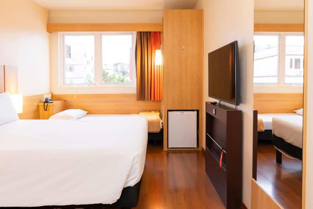 Um quarto no ibis Porto Alegre Moinhos de Vento com cama de casal, TV, frigobar, janela com cortinas laranja e piso de madeira.