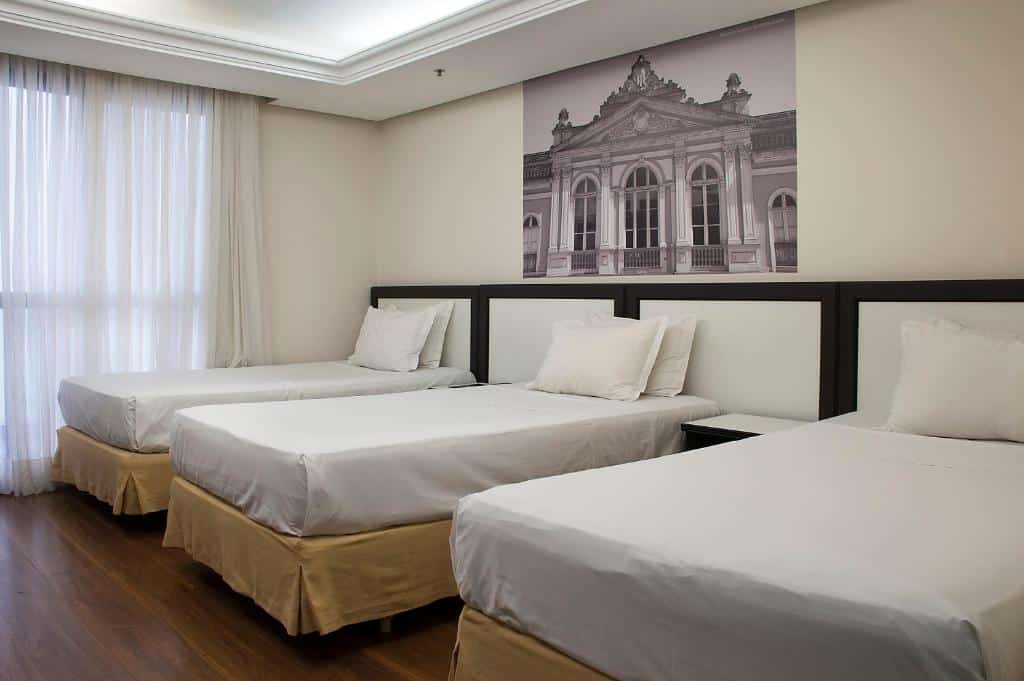 Quarto no Master Grande Hotel com três camas de solteiro, roupa de cama branca, uma grande janela com cortinas e uma foto arquitetônica emoldurada acima das camas.