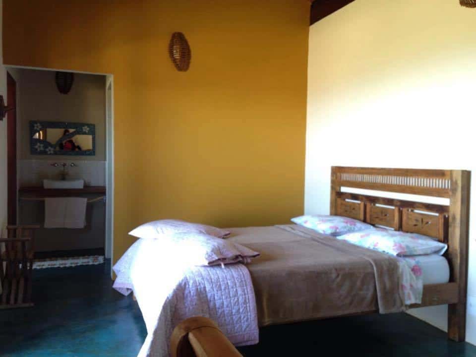 Um quarto na Pousada Encantos do Cerrado com uma cama de madeira, roupa de cama floral, cobertor castanho, parede com detalhes em amarelo e uma porta aberta para o banheiro.