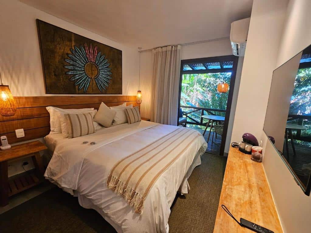 Uma cama bem arrumada em um quarto de hotel com um grande quadro, escrivaninha, TV e uma varanda com vista para a vegetação, em um quarto da Pousada e Mergulho Jamanta.